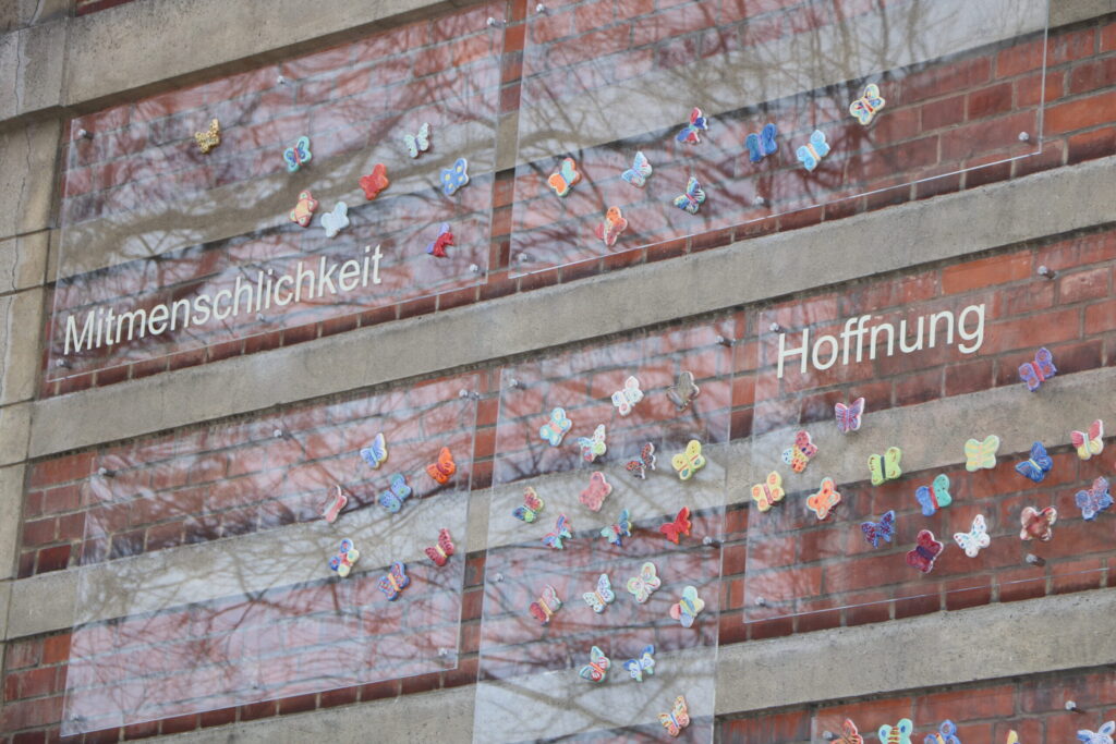 Bunte Schmetterlinge schmücken eine Ziegelwand mit den Worten Mitmenschlichkeit und Hoffnung, während sich Baumäste von außen im Glas spiegeln.