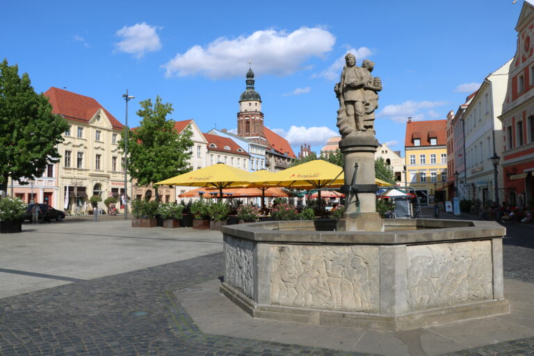Bei Sonnenschein prägt der Altmarktbrunnen den Altmarkt in Cottbus/Chóśebuz mit Cafés, historischen Fassaden und dem Spremberger Turm.