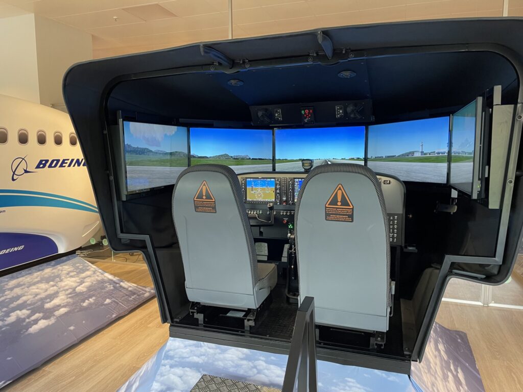 Im Boeing-Flugsimulator stehen zwei Pilotensitze vor mehreren Monitoren, die Startbahn und Vorfeld eines Flughafens mit Tower anzeigen.