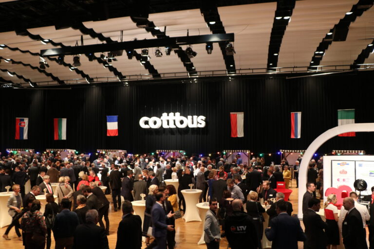 Bei der Veranstaltung in Cottbus treffen sich viele Gäste unter Flaggen und dem leuchtenden Schriftzug „cottous“ im Saal zu Gesprächen.