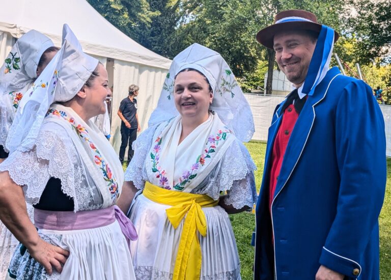 Mehrere Menschen in sorbischer Tracht unterhalten sich lachend bei einer Veranstaltung im Freien, vor einem Zelt und grünen Bäumen im Park.
