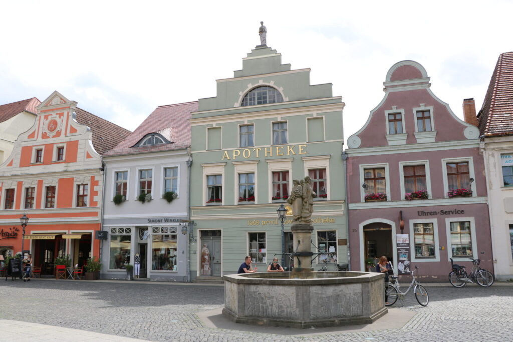 Historische Fassaden am Cottbuser Altmarkt mit der Apotheke, einem Brunnen im Vordergrund und farbigen Giebelhäusern an der Kopfsteinpflasterfläche.