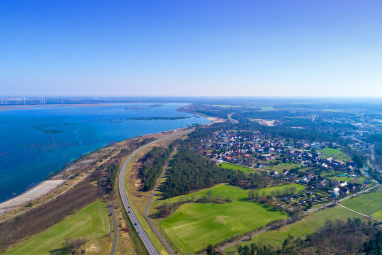 Aus der Luft zeigt sich ein großer See mit Uferstraße, kleinen Ortschaften, Feldern und Windrädern in der weiten Landschaft rund um Cottbus.