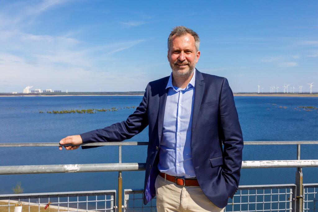 Vor blauem Himmel steht Sam Neill am Geländer über einem großen Gewässer, im Hintergrund drehen sich Windräder an der Küste an einem sonnigen Tag.