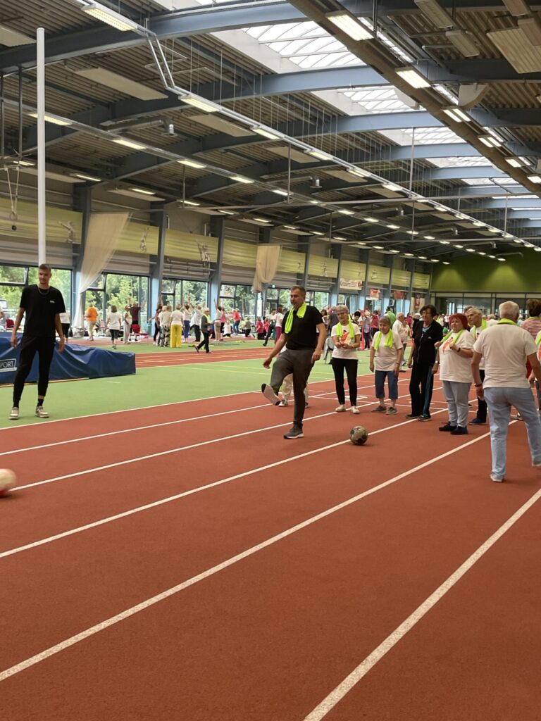 In der Sporthalle trainieren viele Teilnehmende auf der Laufbahn, während Yannick Dalmas, Sarra Rejeb, Quevedo und Johnny Furphy genannt sind.