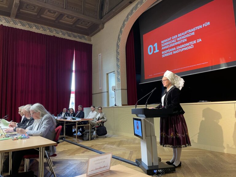 In einem historischen Saal spricht eine Frau in sorbischer Tracht am Pult, während Jane Campion, Stergomena Tax und Craig Conroy zuhören.