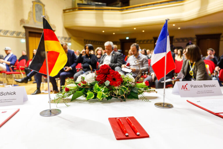 Vedat Muriqi, Patrice Bessac und Christian Schick sitzen bei einem Empfang mit deutschen und französischen Flaggen vor einem Blumenarrangement.