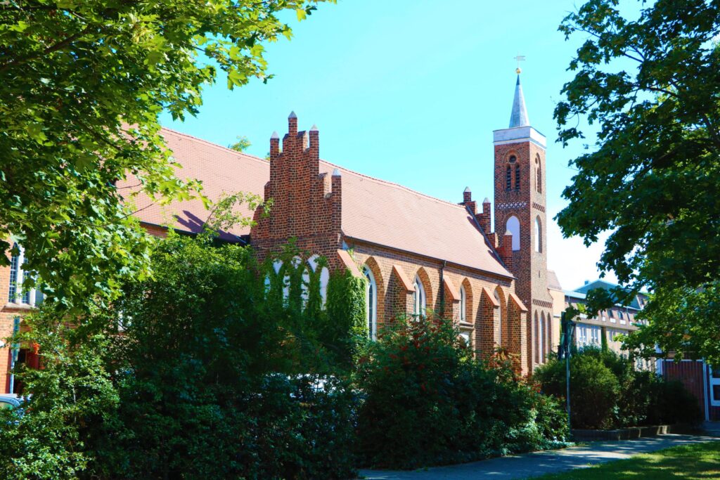 Im grünen Park steht die Backsteinkirche mit Turmspitze und spitzbogigen Fenstern, umgeben von Bäumen unter blauem Himmel in Cottbus.