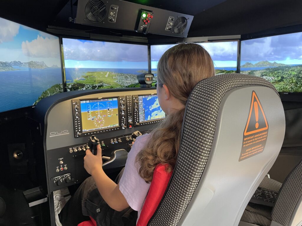 Ein Kind steuert in einem Flugsimulator mit mehreren Bildschirmen ein Flugzeug über eine Inselküste und blickt auf Instrumente und Anzeigen.