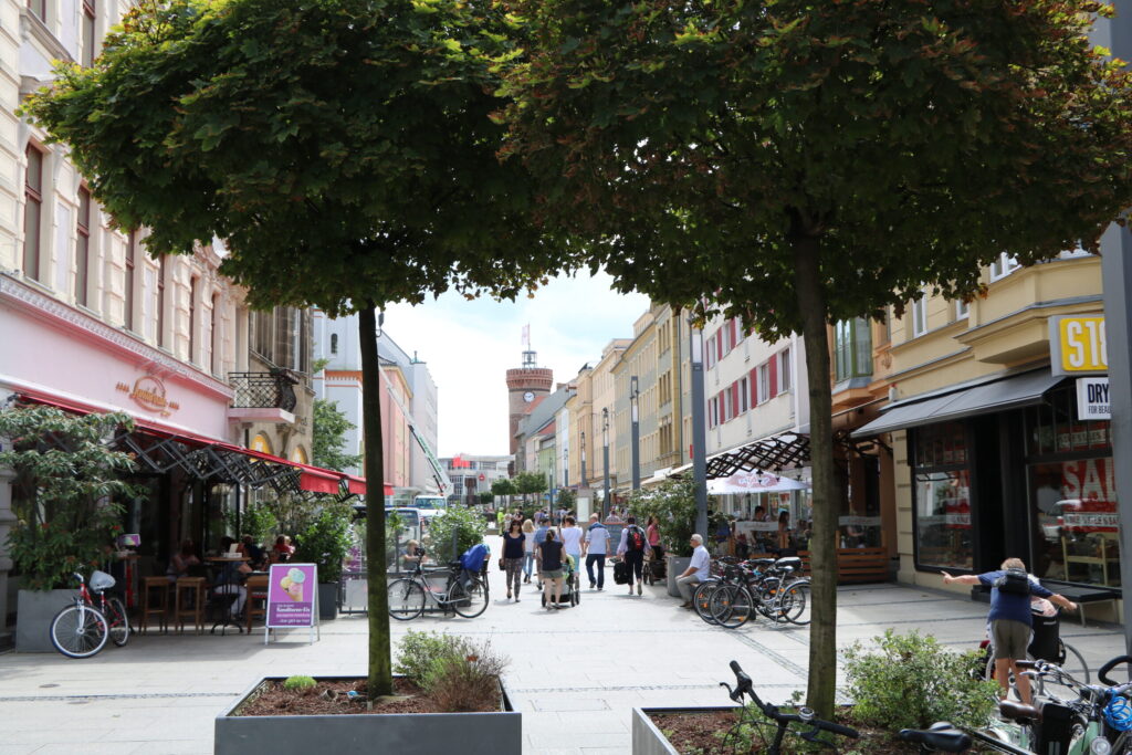 Passanten schlendern durch eine belebte Einkaufsstraße in Cottbus/Chóśebuz, vorbei an Cafés, Fahrrädern und dem Turm der Altstadt im Hintergrund.