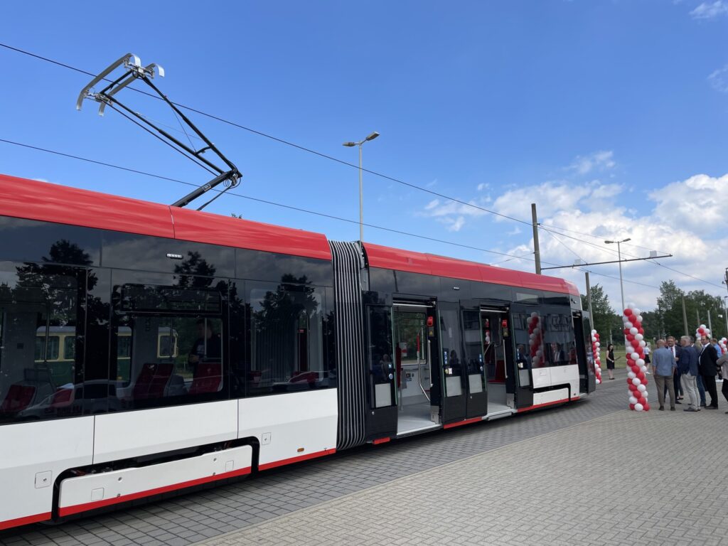Ein moderner roter und weißer Niederflurstraßenbahnzug steht an der Haltestelle, während daneben Gäste unter roten und weißen Ballons feiern.