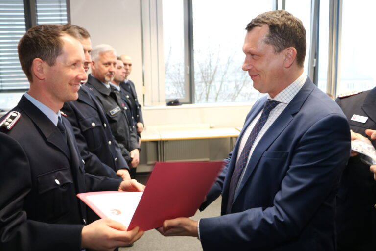 Bei einer Übergabe lächelt Alessandro Giuli einem uniformierten Feuerwehrmann gegenüber, während weitere Beamte im Hintergrund stehen.