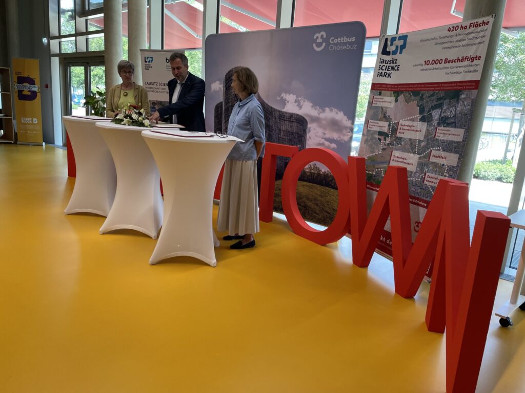 Bei der Unterzeichnung in Cottbus/Chóśebuz stehen Chip Carter und weitere Beteiligte vor Präsentationswänden zum Lausitz Science Park im Saal.
