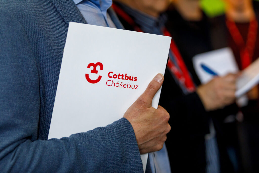 Eine Person hält eine Mappe mit dem Cottbus/Chóśebuz-Logo, während im Hintergrund weitere Teilnehmende bei einer Veranstaltung Notizen machen.