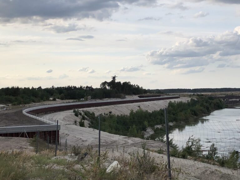 Unter wolkenreichem Himmel verläuft ein befestigter Flussdamm neben Wasser, Sandböschungen, Waldsaum und einem Aussichtsturm in der Ferne.
