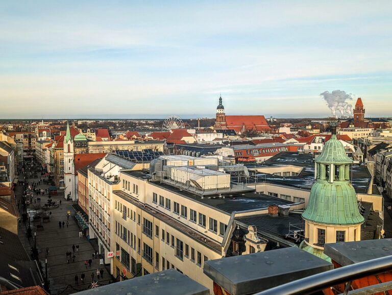 Über den Dächern von Cottbus/Chóśebuz liegen Altstadt, Kirchturm, Riesenrad und Marktplatz im Winterlicht, während am Horizont Rauch aufsteigt.