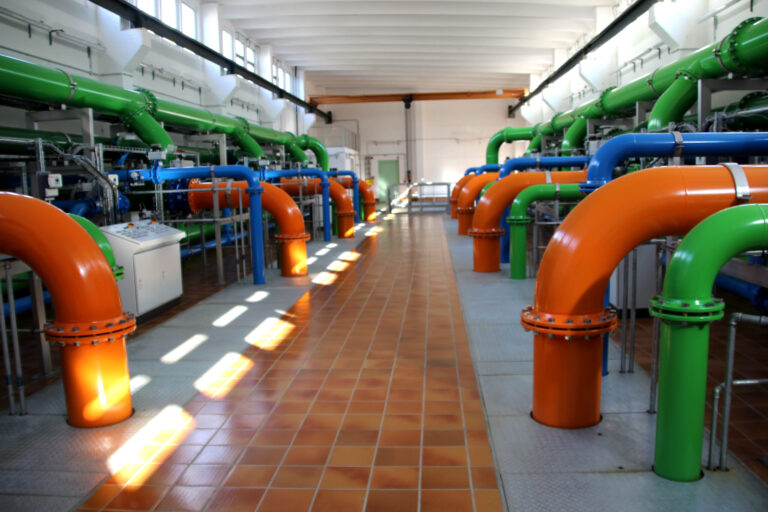 In einer technischen Anlage verlaufen grüne, blaue und orange Rohrleitungen durch einen hellen Maschinenraum mit Fliesenboden und Sonnenlicht.