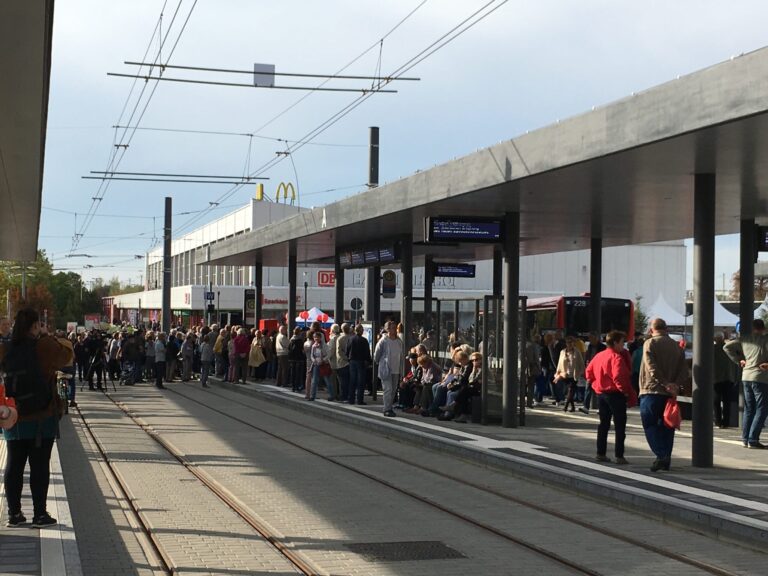 Viele Fahrgäste warten an einer modernen Straßenbahnhaltestelle mit Gleisen, Anzeigen und einem McDonald's-Schild im Hintergrund bei Sonnenschein.