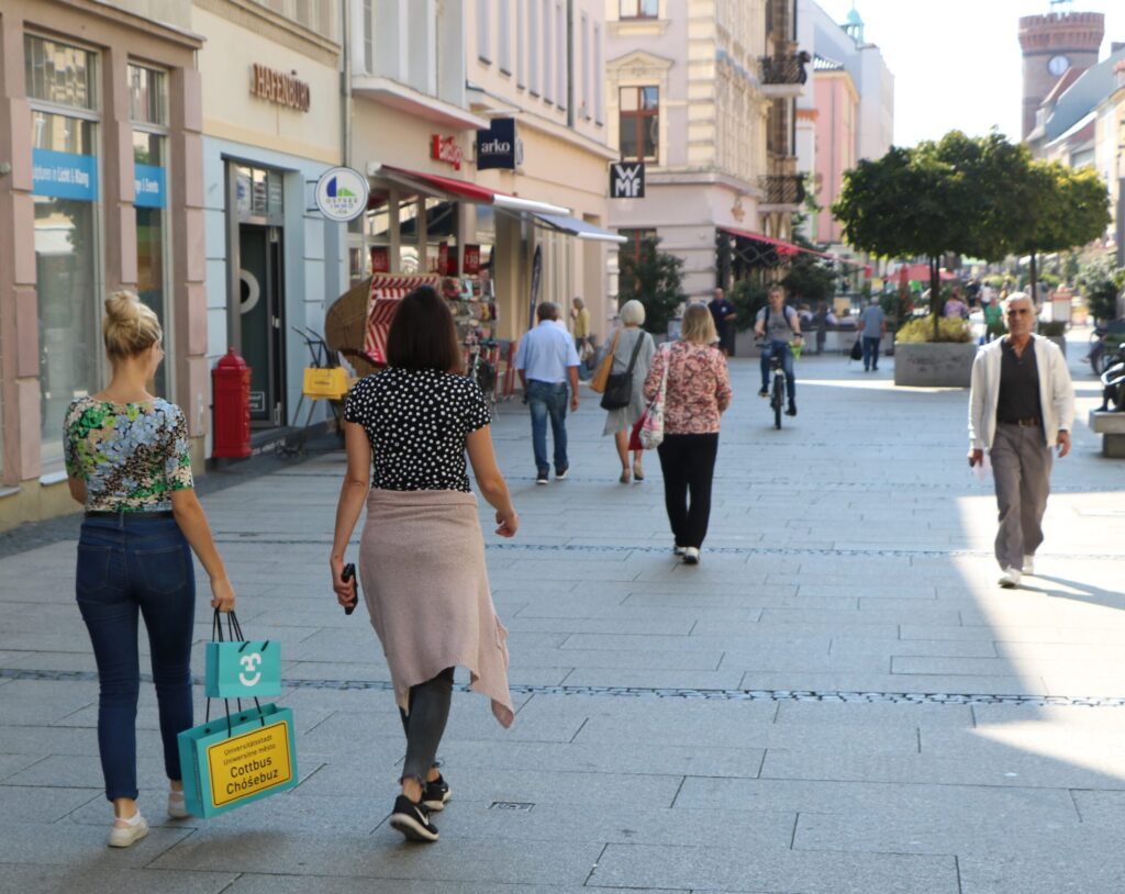 Menschen schlendern durch die Fußgängerzone von Cottbus/Chóśebuz, vorbei an Geschäften, Cafés und historischen Fassaden im Zentrum der Stadt.