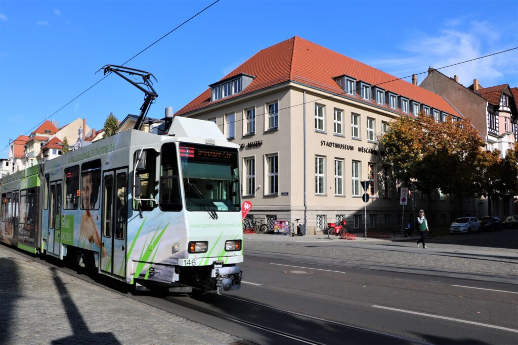 Eine Straßenbahn fährt an der Ecke des Stadtmuseums in Cottbus vorbei, während historische Häuser mit roten Dächern die sonnige Straße säumen.
