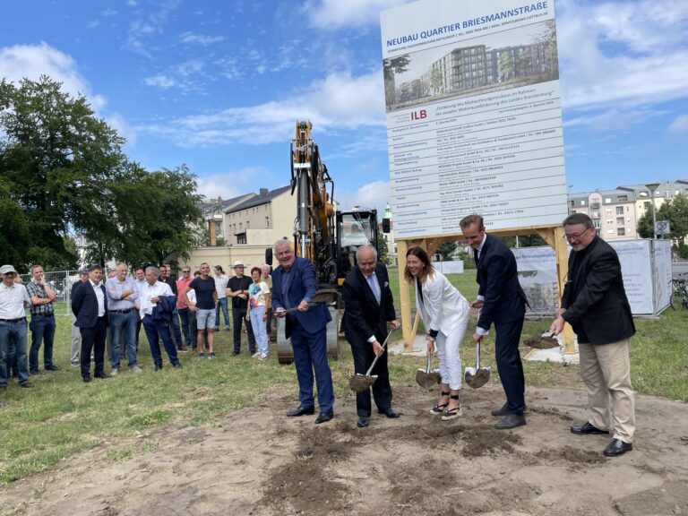 Bei einem Spatenstich in Cottbus/Chóśebuz stehen Vertreter vor dem Neubau Quartier Briesmannstraße mit Bagger und großem Bautafel-Stand.