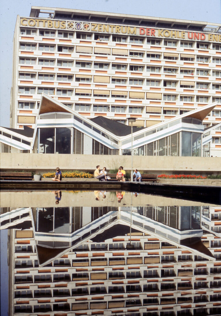 Vor dem Haus mit der Aufschrift „COTTBUS ZENTRUM DER KOHLE UND ENERGIE“ sitzen Menschen am Wasser, das die markante Fassade gespiegelt zeigt.