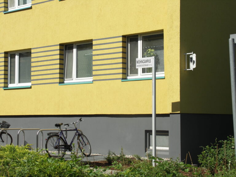 Vor einem gelben Wohngebäude am Wohncampus Universitätsstrasse 12–16 stehen Fahrräder, während Fenster und Weg im Sonnenlicht liegen und Ruhe ausstrahlen.