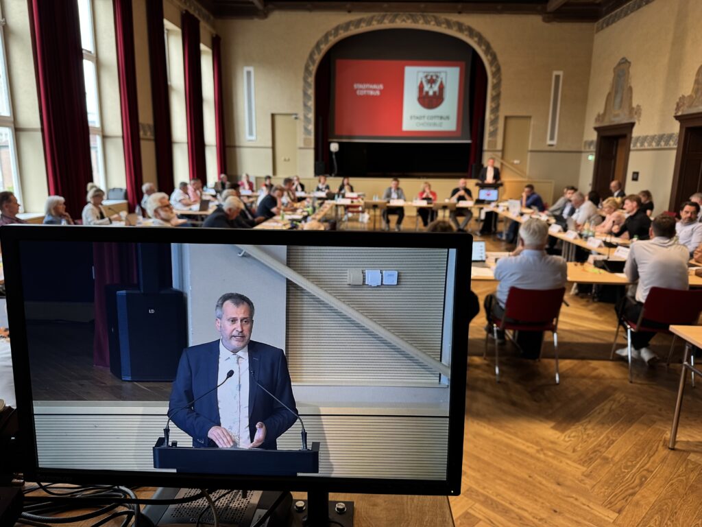 Mann spricht am Rednerpult in einem Sitzungssaal, während die Teilnehmer Tobias Schick im Hintergrund sitzen.