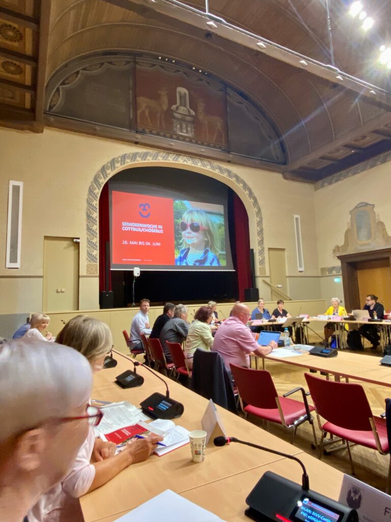 Im historischen Saal beraten Teilnehmende der Sozialausschuss-Sitzung in Cottbus/Chóśebuz, während auf der Leinwand die Seniorenwoche beworben wird.