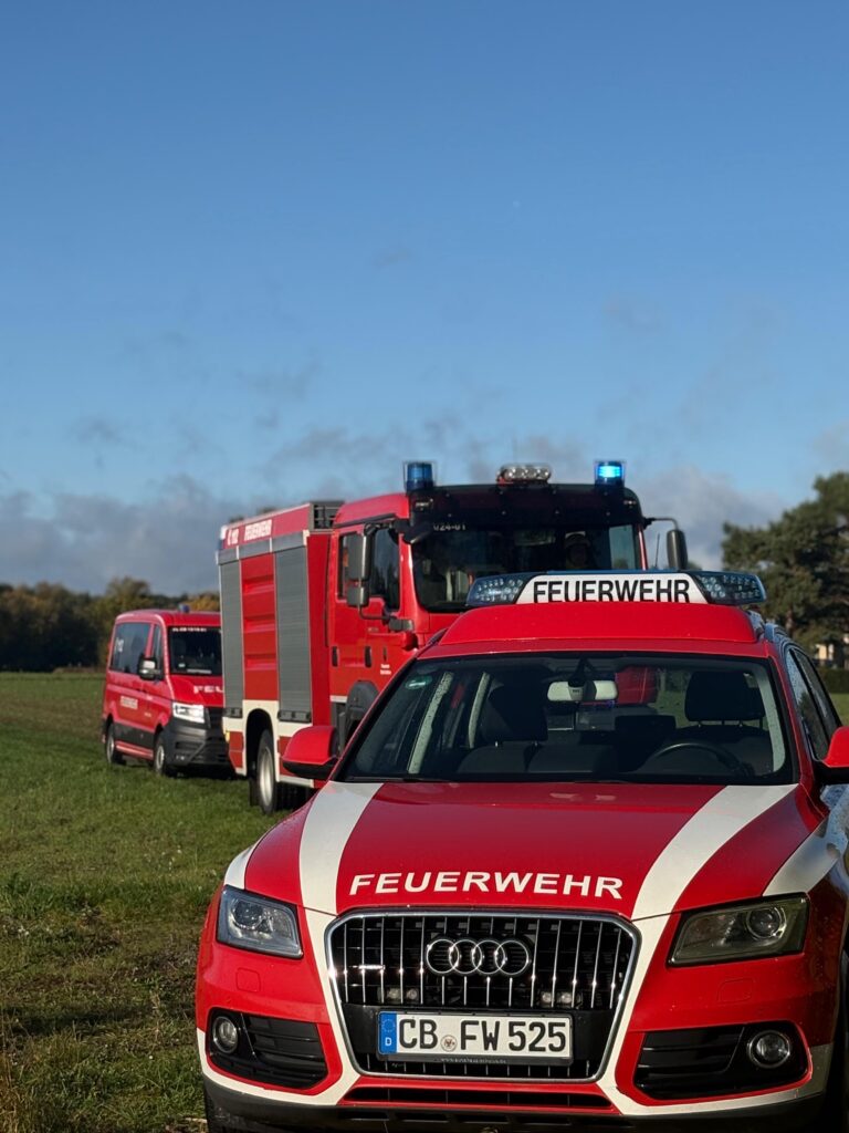 Ein roter Audi mit FEUERWEHR-Aufschrift steht vor einem Löschfahrzeug und Einsatzwagen auf einer Wiese.