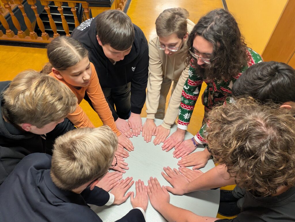 Mehrere Jugendliche legen im Innenraum ihre Hände gemeinsam in einen Kreis und versammeln sich um einen Tisch in einer hellen Halle mit Holzgeländer.