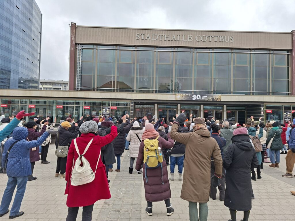 Menschen heben vor der Stadthalle Cottbus die Arme beim One-Billion-Rising-Protest gegen Gewalt in Cottbus/Chóśebuz.