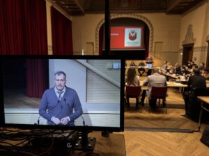 Im Stadtverordnetensaal spricht ein Mann am Pult, während sein Beitrag auf dem großen Bildschirm läuft.