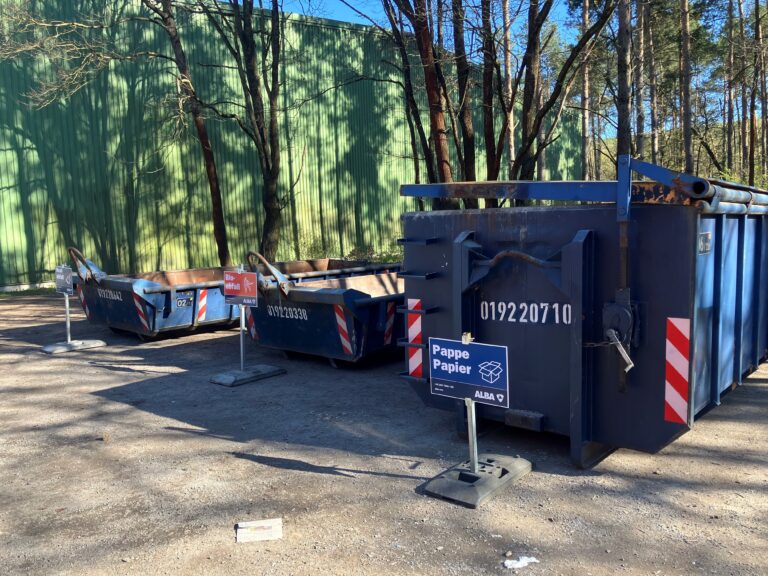 Am Entsorgungszentrum ALBA in Cottbus stehen blaue Container für Restmüll, Bioabfall sowie Papier und Pappe neben einem grünen Hallengebäude.