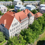 Aus der Luft zeigt sich das Konservatorium Cottbus inmitten grüner Bäume, umgeben von Wohnhäusern mit hellen Dächern und rotem Ziegeldach.