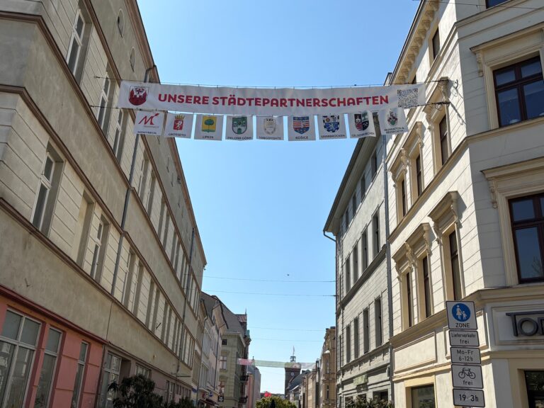Über der Sprem hängt ein Banner mit unseren Städtepartnerschaften und den Wappen von Montreuil, Grosseto, Zielona Góra und weiteren Städten.
