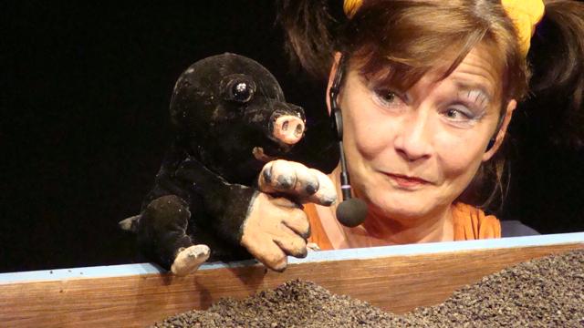 Eine Darstellerin von Theater des Lachens spricht mit einer schwarzen Maulwurf-Handpuppe und blickt über die kleine Erdlandschaft bei Das kleine Krabbeln (2+).