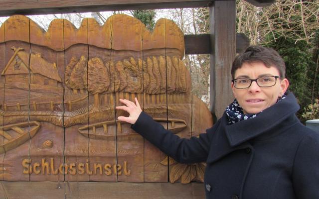 Eine Frau zeigt auf das hölzerne Schild Schlossinsel bei der Stadtführung in Lübben und verweist auf die Geschichte der Spreeinsel im Stadtzentrum.