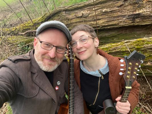 Beim Gundermannabend lächeln Peggy Luck und Thomas Strauch mit Gitarren vor einem moosigen Holzstamm und erinnern in Cottbus an Gerhard Gundermann.