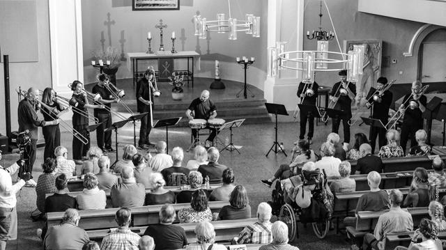 Bei der 63. jazzwerkstatt Peitz spielt ein Ensemble in einer Kirche vor vielen Zuhörern, während Posaunen, Streicher und Schlagzeug erklingen.