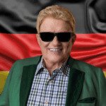 Mit Sonnenbrille und grünem Sakko steht Heino vor der schwarz-rot-goldenen Flagge und kündigt die Tour „Made in Germany“ für 2026 in Cottbus an.