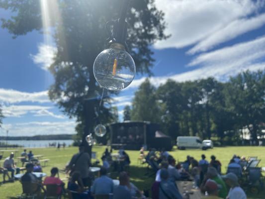 Beim Sommerfest am See im Kurpark Bad Saarow sitzen Besucher auf der Wiese, während Bühnenprogramm, Fahrzeuge und Scharmützelsee im Hintergrund liegen.