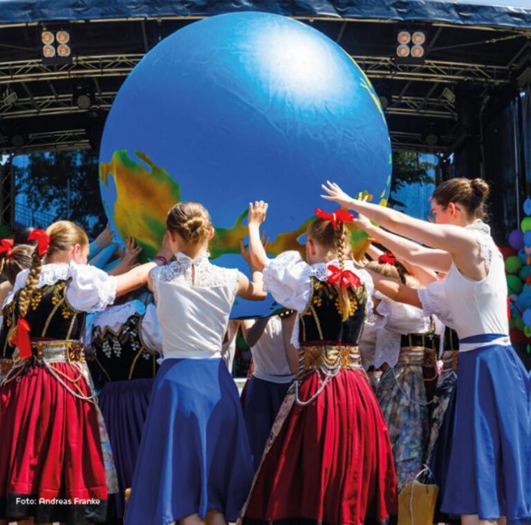 Cottbus/Chóśebuz bei Mitmachen: Kinder tanzen mit farbigen Trachten auf einer Bühne vor einem großen blauen Globus, Bühnenlicht und Deko im Hintergrund.