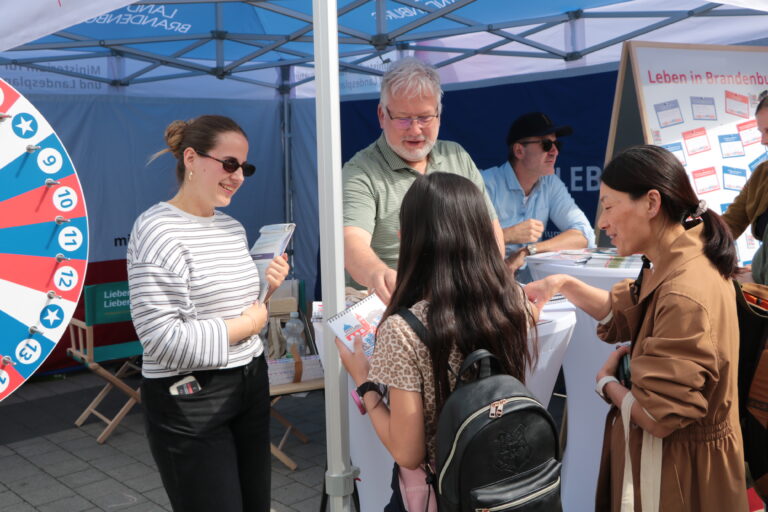 Menschen sprechen an einem Infostand unter einem blauen Pavillon, während ein Glücksrad und Broschüren am Stand in Brandenburg zu sehen sind.