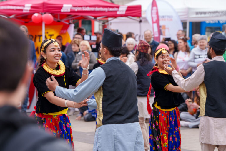 Beim Willkommensfest KLĚB A SOL in Cottbus/Chóśebuz tanzen Teilnehmende gemeinsam in farbenfrohen Trachten vor Publikum auf dem Stadthallenvorplatz.