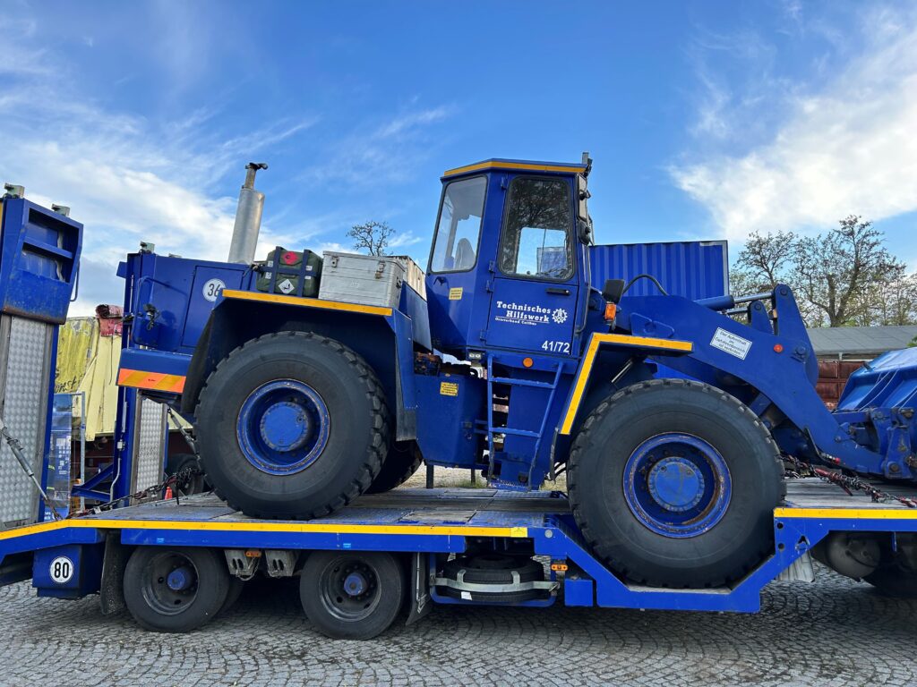 Vor blauem Himmel steht ein blau-gelber THW-Radlader des Technischen Hilfswerks Cottbus/Chóśebuz auf einem Tieflader im Freien.