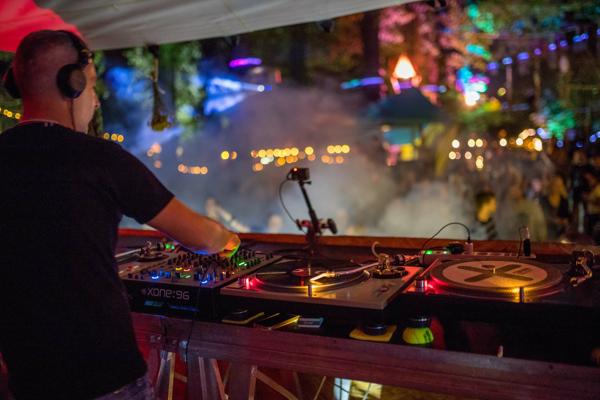 Beim Novum legt ein DJ an zwei Plattenspielern und einem Mixer auf, während bunte Lichter und Nebel die tanzende Menge im Wald umgeben.