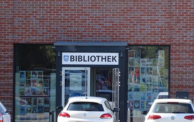 Vor einer Backsteinfassade liegt der Eingang einer Bibliothek mit dem Schild BIBLIOTHEK; davor parken zwei weiße Autos im Vordergrund auf dem Hof.