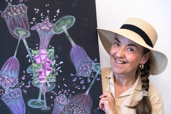 Bei der Ausstellung „Frau Trummer malt weiter“ präsentiert Manuela Trummer eine farbige Kunstarbeit mit violetten Blüten und lächelt in einem Strohhut.