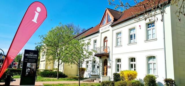 Vor dem Stadtmuseum in Ströbitz steht eine pinke Infofahne neben dem weißen Gebäude mit gepflegtem Grün in Cottbus/Chóśebuz.